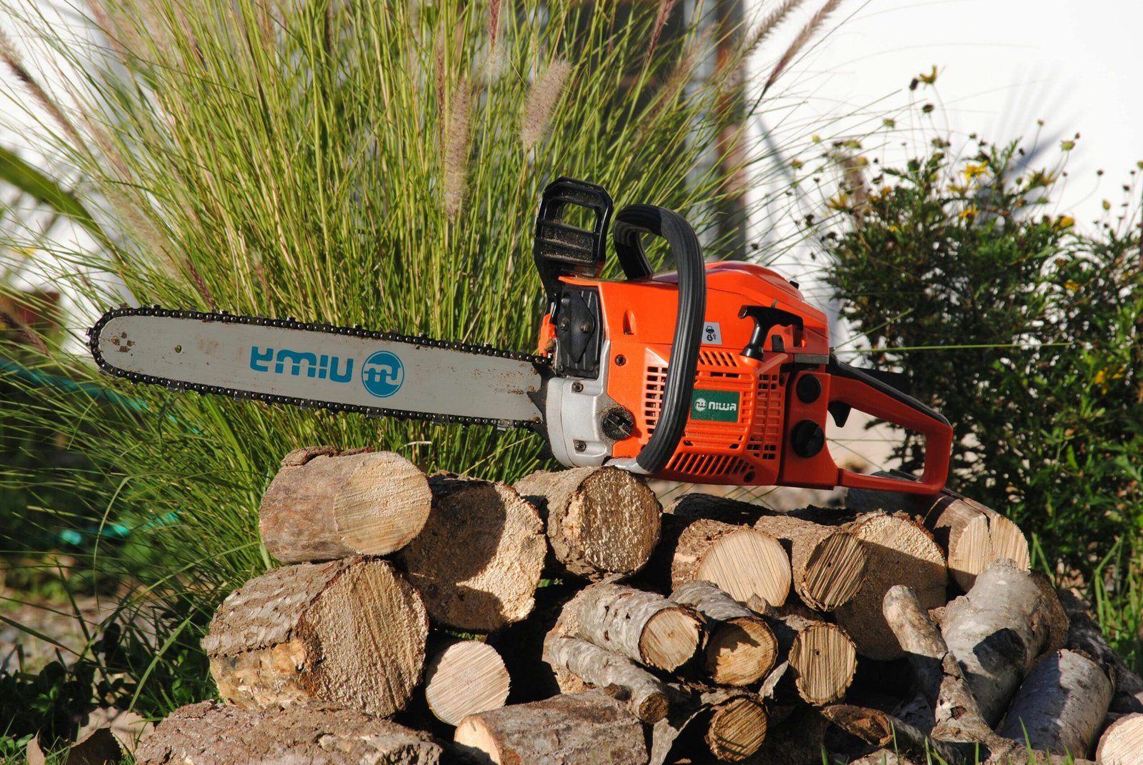 Home Bright orange chainsaw resting on a woodpile outdoors, surrounded by greenery.
